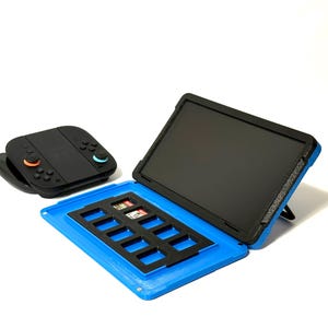 Peut inclure: Une console de jeu portable noire et un étui bleu et noir avec un écran et un rangement pour cartes SD. L'écran est soutenu par un support. La console a des boutons orange et bleus.