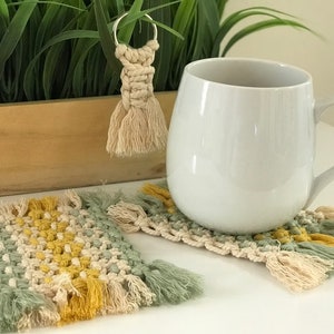 May include: Two crocheted coasters in green, yellow, and cream with fringe. A white ceramic mug sits on one of the coasters. A small crocheted tassel keychain hangs from a ring.