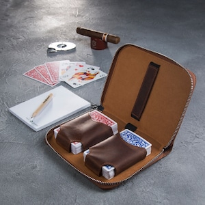 Puede incluir: Un estuche con cremallera de cuero marr&oacute;n con dos barajas de cartas en fundas de cuero marr&oacute;n. Tambi&eacute;n hay un cigarro, un cortador, naipes, un bloc de notas y un l&aacute;piz. El estuche est&aacute; abierto, revelando un interior beige.
