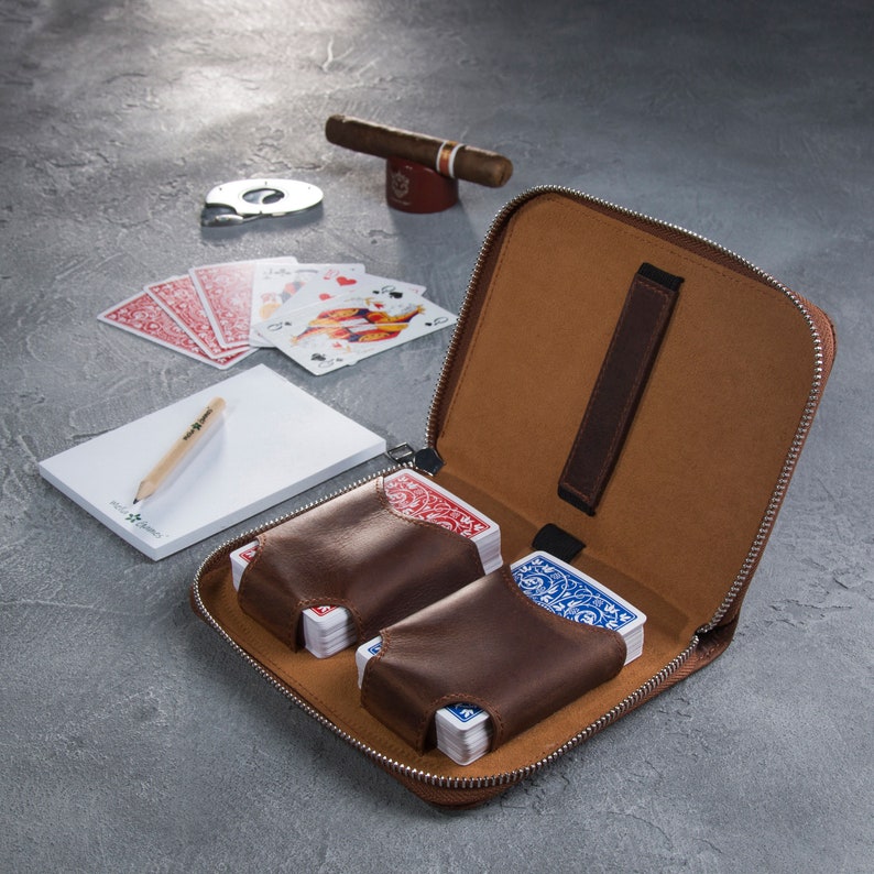 Puede incluir: Un estuche con cremallera de cuero marr&oacute;n con dos barajas de cartas en fundas de cuero marr&oacute;n. Tambi&eacute;n hay un cigarro, un cortador, naipes, un bloc de notas y un l&aacute;piz. El estuche est&aacute; abierto, revelando un interior beige.