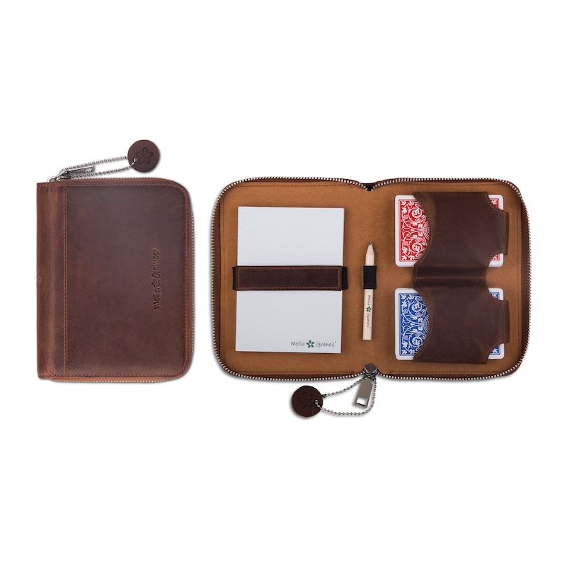 Puede incluir: Un estuche de cuero marr&oacute;n con cremallera, abierto para revelar un bloc de notas, naipes y un l&aacute;piz. El estuche tiene una leng&uuml;eta de cuero a juego y una cremallera met&aacute;lica. El bloc de notas se sujeta con una correa de cuero marr&oacute;n. Las cartas son rojas y azules.