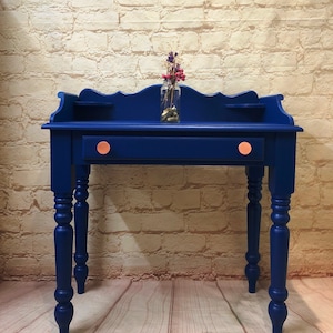 May include: A dark blue wooden desk with a single drawer and turned legs. The desk has two small shelves on the back and copper drawer pulls. A small glass vase with dried flowers sits on the top of the desk.