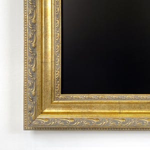Può includere: Una cornice rettangolare con finitura oro metallizzato e un motivo a rilievo ornato con disegni di foglie e un bordo interno perlato. La cornice circonda un pannello centrale rettangolare nero opaco, liscio e uniforme.