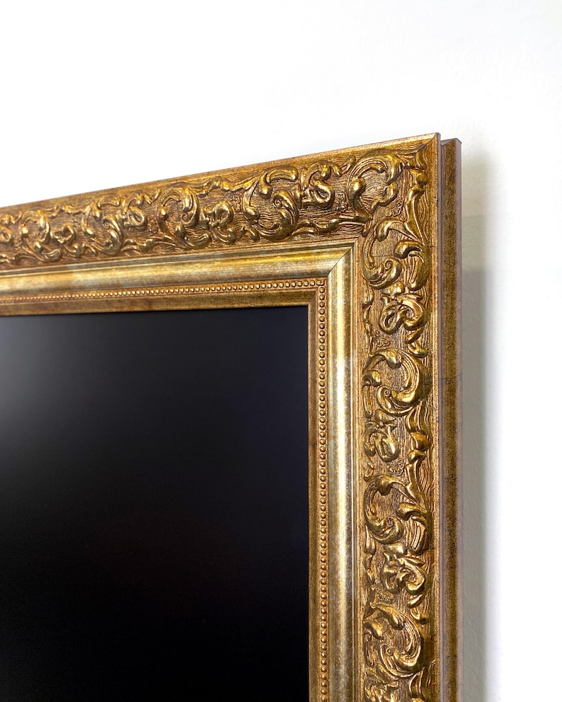 May include: Ornate gold picture frame with a black interior. The frame features detailed relief carvings and a metallic finish. The corner of the frame is visible against a white background, showcasing the intricate design and texture.