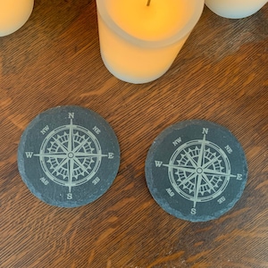 May include: Two black slate coasters with a compass design engraved on them. The compass features the cardinal directions N, S, E, and W, as well as the intermediate directions NW, NE, SW, and SE.