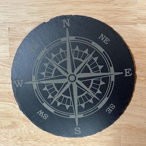 May include: A round, dark gray slate coaster with a compass rose design etched into the surface. The compass points are labeled N, NE, E, SE, S, SW, and W. The coaster is approximately 4 inches in diameter.