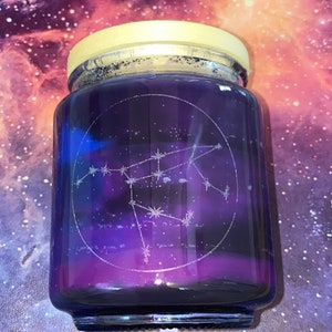 May include: A glass jar candle with a celestial theme. The candle has a gradient of purple and blue hues, with a constellation design etched on the glass. The lid is a light yellow color.