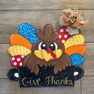 Pode incluir: Um peru de madeira pintado com penas coloridas e uma panela preta com o texto "Give Thanks". O peru está olhando para o espectador com uma expressão de surpresa.