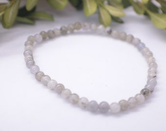 May include: A delicate bracelet made of small, round, light gray beads. The beads have a subtle shimmer and are strung together on a clear elastic cord.