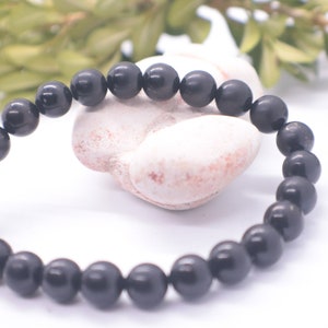 May include: A black beaded bracelet made of round, smooth stones. The bracelet is lying on a white surface with a white rock behind it.