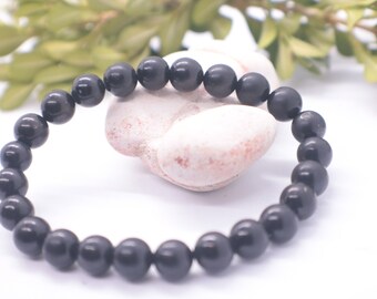 May include: A black beaded bracelet made of round, smooth stones. The bracelet is lying on a white surface with a white rock behind it.