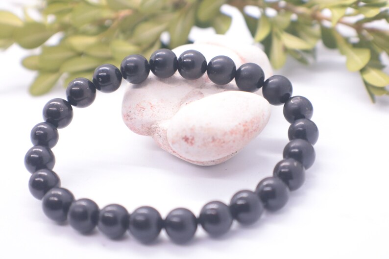 May include: A black beaded bracelet made of round, smooth stones. The bracelet is lying on a white surface with a white rock behind it.