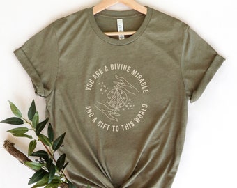 May include: A green t-shirt with a white graphic that says "You are a divine miracle and a gift to this world". The graphic features a stylized tree with stars and hands holding it.