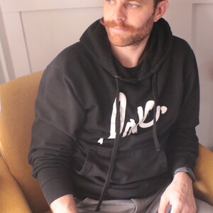 May include: A man wearing a black hoodie with a white logo that says "Jaker" is sitting in a yellow armchair. He is holding a black mug with a white design.