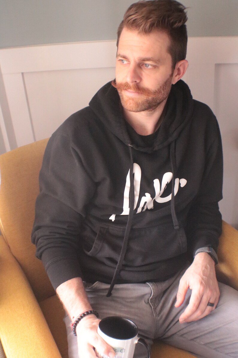 May include: A man wearing a black hoodie with a white logo that says "Jaker" is sitting in a yellow armchair. He is holding a black mug with a white design.