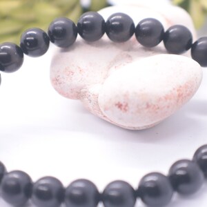 May include: A black beaded bracelet made of round, smooth stones. The bracelet is lying on a white surface.