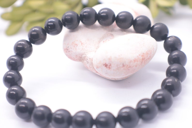 May include: A black beaded bracelet made of round, smooth stones. The bracelet is lying on a white surface.