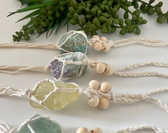 Puede incluir: Cuatro piedras de cristal envueltas en macramé con cuentas de madera. Las piedras son verdes, moradas, amarillas y una mezcla de verde y morado.