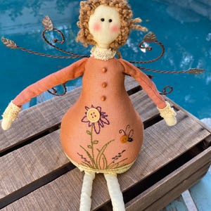 May include: A handmade doll with a peach-colored dress and curly brown hair. The doll has a floral design with a button and a bee. It has wire wings and white legs. The doll is sitting on a wooden crate.