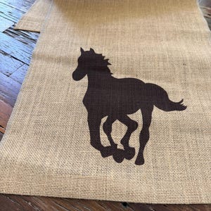 May include: A tan burlap table runner featuring a dark brown silhouette of a running horse. The horse is depicted in a dynamic pose, with its legs extended. The runner is made of a coarse, woven fabric.