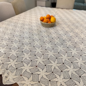 May include: A cream-colored, crocheted tablecloth with a geometric pattern covers a wooden table. A small woven basket filled with oranges and a lemon sits in the center. The tablecloth's edge has a scalloped design.