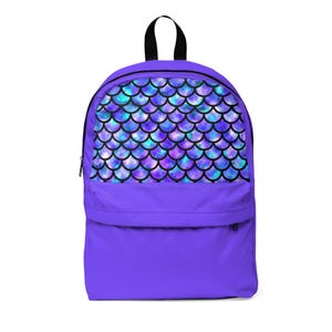 Puede incluir: Una mochila morada con asa y cremallera negras. La sección superior presenta un patrón de escamas de sirena en tonos azules, morados y turquesas. La mochila tiene un bolsillo frontal y está diseñada para transportar objetos.
