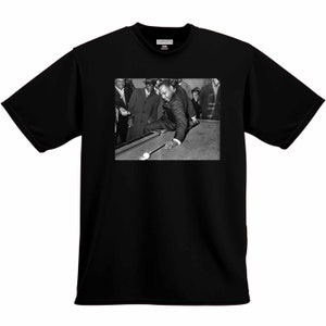 May include: Black and white photo of a man playing pool. He is wearing a suit and tie and is leaning over the table to take a shot. There are other people in the background watching him play.