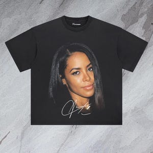 May include: Black t-shirt featuring a printed image of a woman with long dark hair and a white signature. The signature reads "Aaliyah".