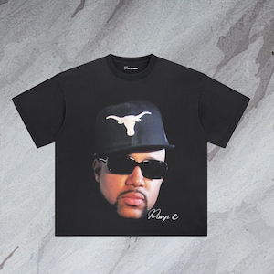 May include: Black t-shirt with a graphic print of a man wearing a black baseball cap with a white Texas Longhorns logo, sunglasses, and a goatee. The text "Pimp C" is printed in white below the graphic.