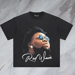 May include: Black t-shirt featuring a portrait of a person wearing star-shaped sunglasses. The image has a signature that says "Rod Wave" in white cursive script. The shirt is a crew neck.