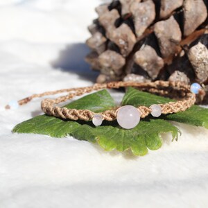 May include: A light pink rose quartz bead and two smaller white opal beads are strung on a brown braided cord bracelet.