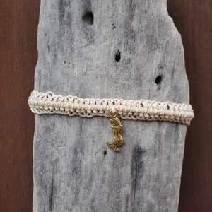 May include: A white macrame cord bracelet with a gold mermaid charm. The bracelet is on a weathered wooden surface.