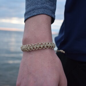 Puede incluir: Primer plano de una muñeca con un brazalete de macramé beige con cierre anudado.