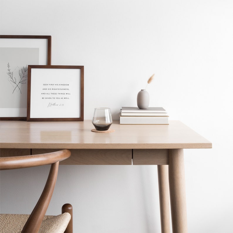 May include: A wooden desk with a light wood finish. A framed print with the text "Seek first his kingdom and his righteousness, and all these things will be given to you as well" is on the desk. A glass of water and a vase with a single dried flower are also on the desk.