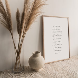 May include: A framed print with a white background and black text. The text reads "Let us then approach God's throne of grace with confidence so that we may receive mercy & find grace to help us in our time of need. Hebrews 4:16". The print is sitting on a white surface with a vase of dried pampas grass and a small beige ceramic vase.