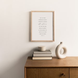 May include: A framed print with a white background and black text. The text reads: "Let us then approach God's throne of grace with confidence so that we may receive mercy & find grace to help us in our time of need. Hebrews 4:16". The print is sitting on a wooden shelf with a stack of books and a white ceramic vase.