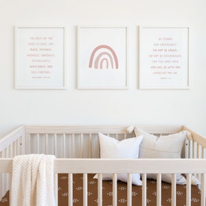 May include: Three framed prints on a white wall above a crib. The left print features a bible verse about the fruit of the spirit. The middle print features a pink and peach rainbow. The right print features a bible verse about being strong and courageous.
