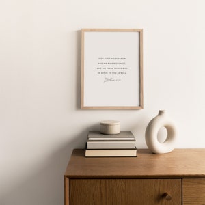 May include: A framed print with a white background and black text that reads "Seek first his kingdom and his righteousness, and all these things will be given to you as well. Matthew 6:33". The print is hanging on a white wall above a wooden dresser with a white vase and a stack of books.