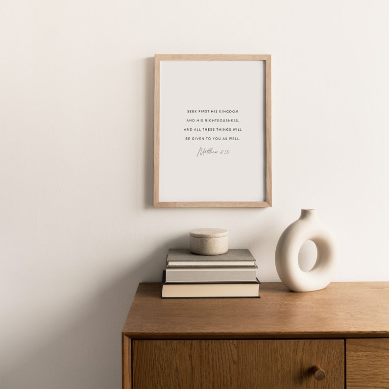 May include: A framed print with a white background and black text that reads "Seek first his kingdom and his righteousness, and all these things will be given to you as well. Matthew 6:33". The print is hanging on a white wall above a wooden dresser with a white vase and a stack of books.