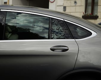 May include: A silver car side profile featuring a rear passenger window decorated with a repeating black bat pattern. The vehicle has a chrome window trim, a body-colored door handle, and tinted glass reflecting the sky and surrounding buildings.