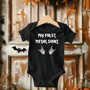 Może przedstawiać: Czarny śpioszek dla niemowlęcia z białym napisem "My First Metal Shirt" i dwoma białymi szkieletowymi rękami wykonującymi gest rock and roll.