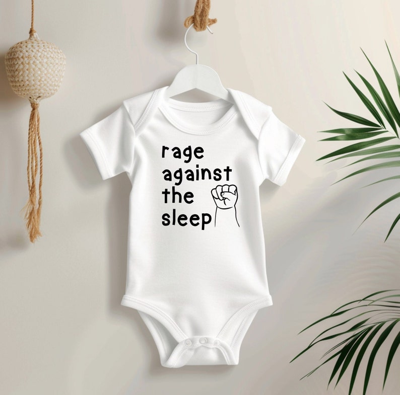 May include: White baby onesie with short sleeves, hanging on a white hanger. The onesie features the text "rage against the sleep" in black, with a black fist graphic. The onesie is made of soft material.