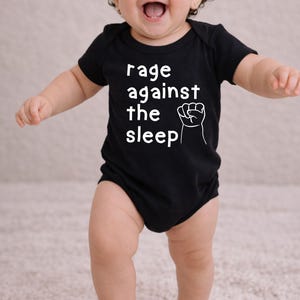 May include: A black baby onesie with the text "rage against the sleep" and a white fist graphic. The baby is smiling and standing on a light-colored rug.