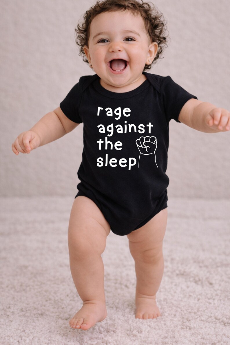 May include: A black baby onesie with the text "rage against the sleep" and a white fist graphic. The baby is smiling and standing on a light-colored rug.