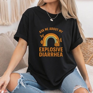 May include: Black t-shirt with a rainbow graphic and the text "Ask Me About My Explosive Diarrhea". The rainbow design includes orange, yellow, and green colors, with butterflies and flowers. The shirt is made of a soft material.
