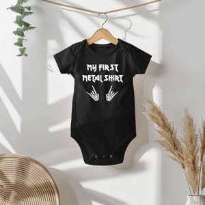 Może przedstawiać: Czarny śpioszek dla niemowlęcia z białym napisem "My First Metal Shirt" i dwoma szkieletowymi rękami wykonującymi znak rock and roll.