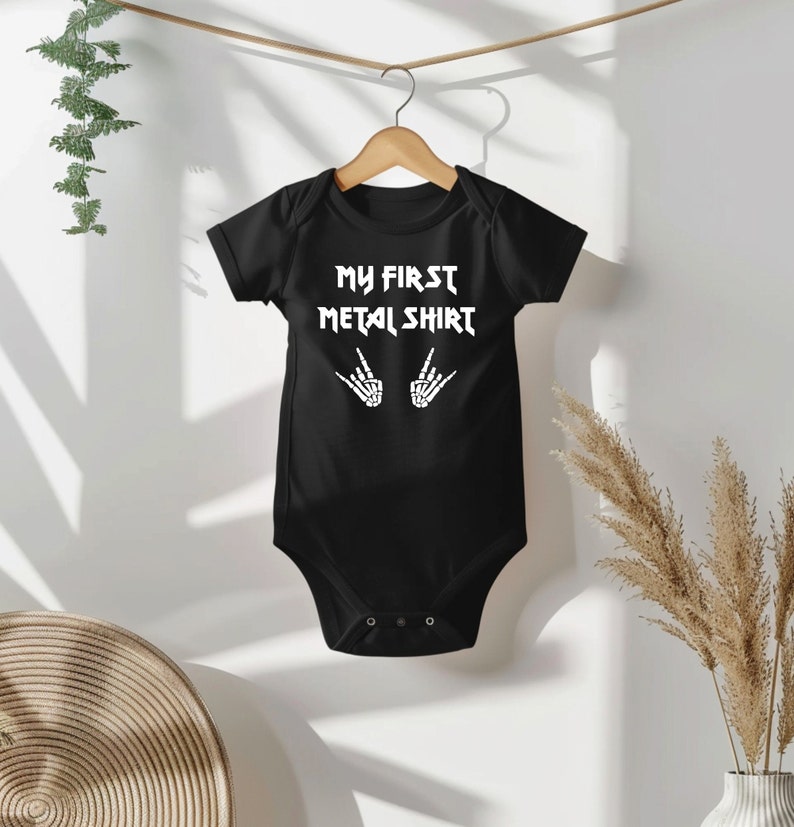 Może przedstawiać: Czarny śpioszek dla niemowlęcia z białym napisem "My First Metal Shirt" i dwoma szkieletowymi rękami wykonującymi znak rock and roll.
