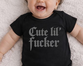 May include: A black baby bodysuit with the text "Cute lil' fucker" in a gothic font. The baby is smiling and has curly hair. The bodysuit is short-sleeved.