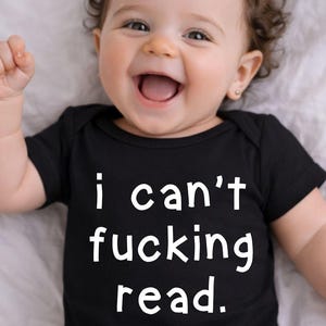 Può includere: Un bambino sorridente indossa un body nero con la scritta bianca "i can't fucking read." Il bambino ha i capelli ricci castani ed è sdraiato su una superficie bianca.