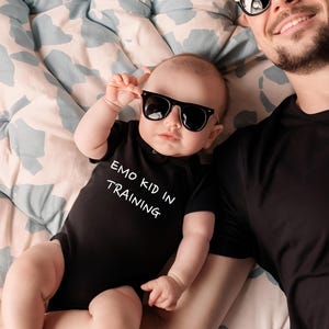 May include: A baby wearing a black onesie with the text "Emo Kid in Training" and black sunglasses. The baby is being held by an adult wearing a black shirt and sunglasses.